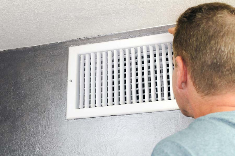 Mature male peering inside an upper wall white grid air duct on a silver wall near a white ceiling. A guy inspecting a heating and cooling air register duct for maintenance and because of air conditioner noise.