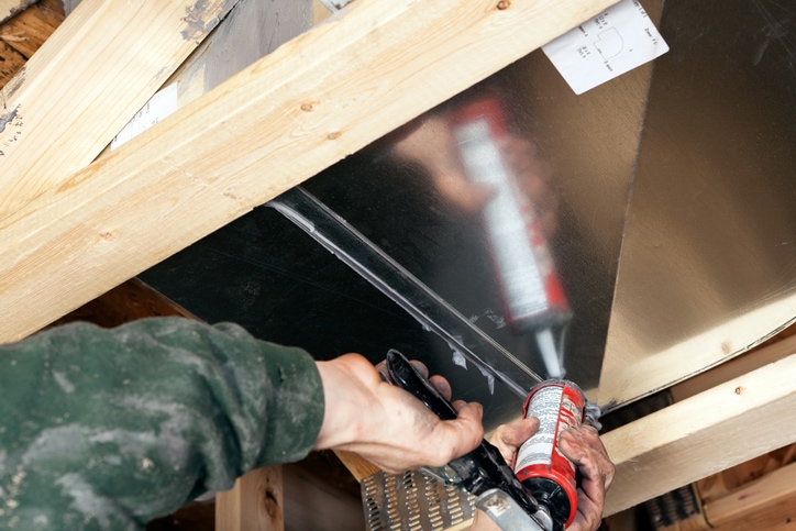 Male hands are using a caulk gun to apply sealant to a house air duct. Caulking ducts prevents air-conditioned or heated air from escaping the duct and prevents ambient air from entering the system. This is a new practice in homebuilding to increase energy efficiency, eventually it may become standard or mandatory.